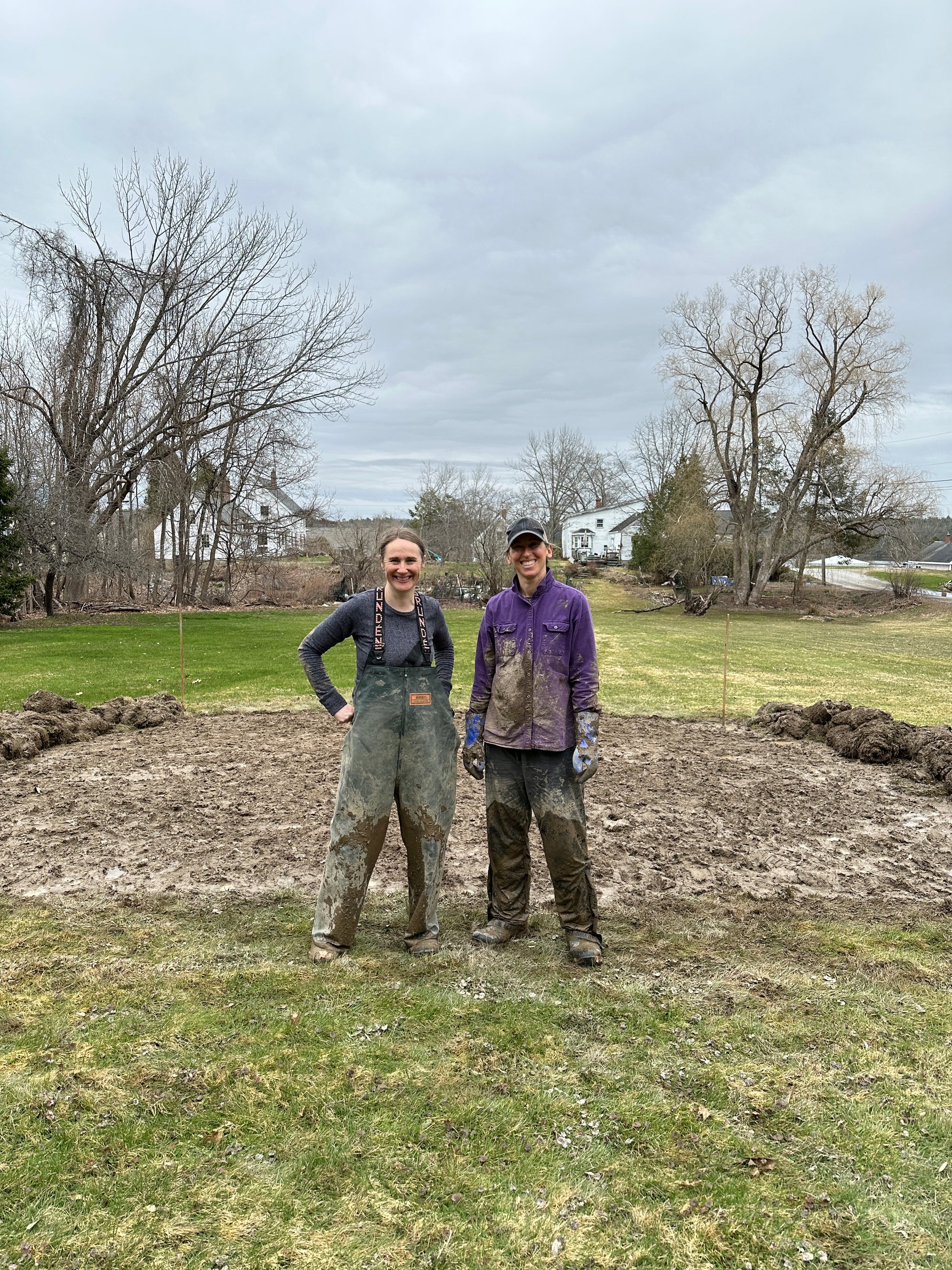 Two women pose in muddy waders in the middle of a field that is being prepped for planting dahlias. 